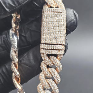 May include: A gold-toned, diamond-studded chain bracelet. The bracelet features a thick curb link design, with each link and the clasp covered in small, sparkling diamonds. The clasp is rectangular and also covered in diamonds. The bracelet is held against a dark background.