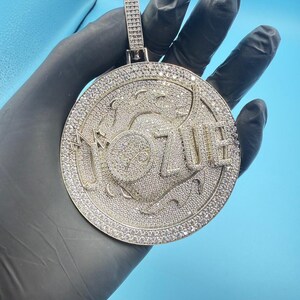 May include: A large, round pendant with the word "OZUE" in raised letters. The pendant is covered in small, sparkling stones, and has a rectangular bail at the top for a chain. The background is a solid light blue.