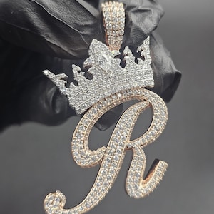 Puede incluir: Un colgante dorado con la letra "R" incrustada con brillantes piedras blancas. Un diseño de corona, también adornado con piedras blancas, corona la letra. El colgante es probablemente una joya, posiblemente un collar o un charm.