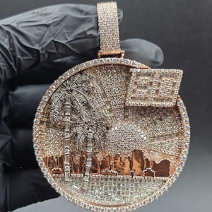May include: A round, rose gold pendant with a detailed cityscape design. The pendant features palm trees, a sunset, and a flag, all encrusted with sparkling gemstones. The bail is also encrusted with gemstones.