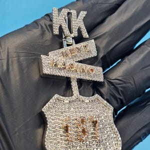 Könnte beinhalten: Ein diamantbesetzter Anhänger mit den Buchstaben "KK" oben, gefolgt von "KEYZ KASINO" in goldfarbenen Buchstaben auf einem rechteckigen Balken. Der Anhänger hat die Form eines Schildes mit den goldenen Zahlen "187".