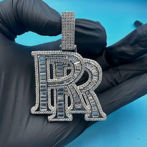 May include: A silver pendant in the shape of the letters "RR" encrusted with small, sparkling stones. The pendant is held against a blue background, showcasing its intricate design and reflective surface. The pendant is a piece of jewelry.