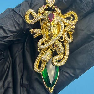 Può includere: Ciondolo dorato con design Medusa, con un volto centrale, serpenti e dettagli di pietre preziose. Ha una grande pietra verde a forma di goccia alla base ed è impreziosito da piccole pietre preziose rosse e trasparenti.