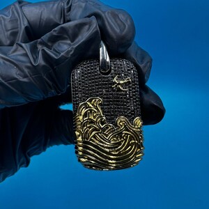 May include: A rectangular pendant with a silver bail. The pendant features a black, textured surface with a gold wave design at the bottom. A small gold emblem is visible near the top. The pendant is held by a gloved hand against a blue background.