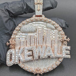 May include: A large, ornate pendant with the word "OKEWA LE" spelled out in sparkling, diamond-like stones. The pendant features a cityscape design and a cross at the top, set in a gold-colored metal.