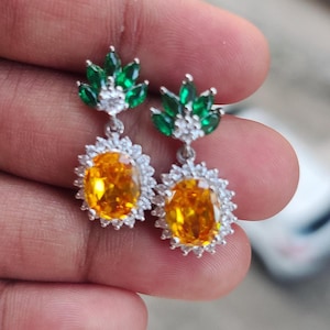 Op de afbeelding: Zilverkleurige oorhangers. Elke oorbel heeft een groep groene, bladvormige stenen aan de bovenkant, een grote ovale gele steen in het midden en een halo van kleine witte stenen.