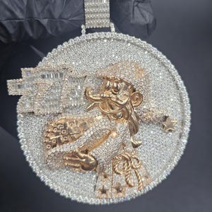 May include: A large, round pendant with a detailed design. The pendant features a central figure in gold, surrounded by a border of small, sparkling stones. The bail is made of rectangular stones. The overall design is intricate and eye-catching.