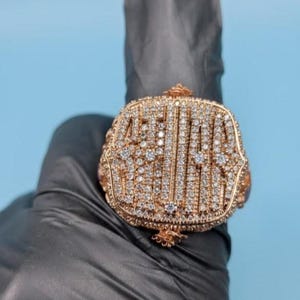 Puede incluir: Un anillo grande, de color dorado, con una cara rectangular, incrustado con numerosas piedras preciosas pequeñas y transparentes. La cara del anillo presenta un diseño ornamentado con las letras "ABIAL" en disposición vertical. El anillo se sostiene sobre un fondo azul claro.