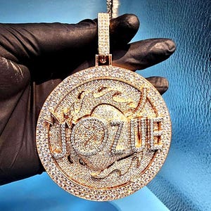 May include: A large, round gold pendant encrusted with numerous small, sparkling gemstones. The pendant features the word "JOZUE" in a stylized font, also outlined with gemstones. The bail is also covered in gemstones. The pendant is suspended from a silver chain.