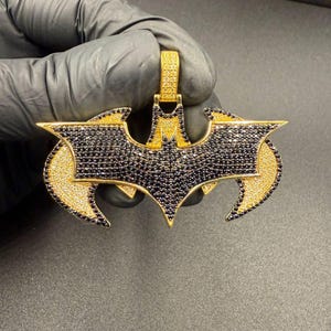 May include: A gold-coloured pendant in the shape of a bat symbol, encrusted with black and yellow gemstones. The pendant is held by a gloved hand against a dark background. The pendant is a piece of jewellery.