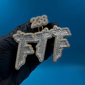 May include: A gold and silver pendant with the letters "FTT" encrusted with small, sparkling stones. The number "238" is above the letters. The pendant is held against a blue background.