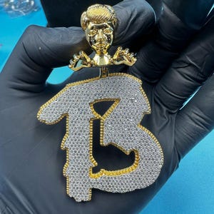 May include: A gold-toned pendant featuring a stylized figure above a large, diamond-studded letter "B". The letter is outlined in gold, and the pendant is held by a gloved hand against a blue background. The pendant is a piece of jewelry.