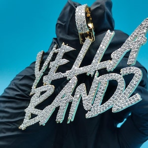 Può includere: Un ciondolo dorato con la scritta "VELLA BANDZ" incastonata con numerose piccole gemme trasparenti. Il ciondolo è tenuto su uno sfondo blu, con un guanto nero parzialmente visibile.