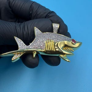 Puede incluir: Un colgante de tiburón dorado, adornado con piedras preciosas claras y rojas. El cuerpo del tiburón está cubierto de pequeñas piedras claras, con un ojo de piedra roja. El colgante se muestra sobre un fondo azul.
