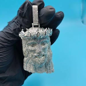 May include: A silver-colored pendant in the shape of a face, possibly Jesus, with a crown of thorns. The pendant is covered in small, sparkling stones. The bail is also encrusted with stones. The background is a solid light blue.