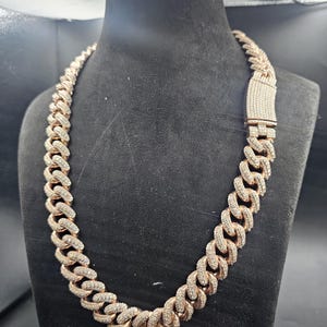May include: A rose gold chain necklace with a cuban link design. The links are encrusted with small, clear gemstones. The necklace is displayed on a black mannequin bust.