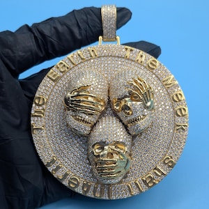 May include: A gold-colored pendant featuring three skull designs, each with a hand covering the eyes, ears, and mouth. The pendant is encrusted with small, sparkling stones and has the text "The Meek Shall Inherit The Earth" around the edge.