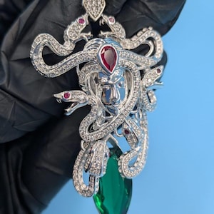 Peut inclure: Pendentif argenté représentant Méduse, orné de serpents, de pierres rouges et de cristaux transparents. Une grosse pierre verte en forme de goutte est suspendue en dessous. Le pendentif est présenté sur fond bleu.