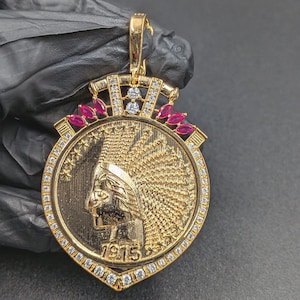 May include: A gold-colored pendant featuring a Native American profile, encircled by a diamond-studded border. The pendant has a crown design with red and white gemstones. The year "1975" is visible.