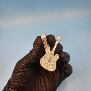 Può includere: Un ciondolo a forma di chitarra tempestato di diamanti con una cauzione abbinata. Il ciondolo è tenuto da una mano guantata su uno sfondo azzurro. Il ciondolo a forma di chitarra è ricoperto di piccole pietre scintillanti e la cauzione ha un design decorativo.