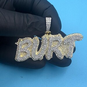 Puede incluir: Un colgante dorado con la palabra "BURT" escrita en cursiva. Las letras están incrustadas con pequeñas piedras blancas brillantes. El colgante se muestra sobre un fondo azul.