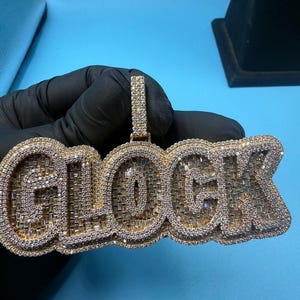 以下が含まれることがあります： 大胆で様式化されたフォントで「GLOCK」と書かれた金色のペンダント。文字はキラキラと輝く透明な宝石で覆われており、豪華なデザインです。ペンダントは、お揃いのベイルから吊り下げられており、着用できます。