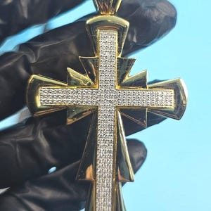 May include: Gold-tone cross pendant with a diamond-like encrusted centre and accents. The cross features a detailed design with a polished finish, showcasing a blend of elegance and religious symbolism. The pendant is suspended from a matching bail.