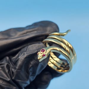 Peut inclure: Une bague serpent dorée avec des yeux en pierres précieuses rouges. La bague est enroulée, avec la tête du serpent en haut. La bague est tenue sur un fond bleu.