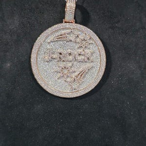 May include: A round pendant necklace with a silver chain. The pendant has a rose gold border and is encrusted with small, sparkling stones. The word "ROCK" is in the center of the pendant, also in rose gold.