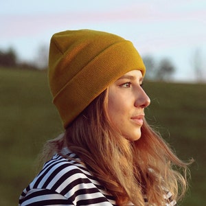 Könnte beinhalten: Eine Frau mit einer gelben Mütze und einem schwarz-weiß gestreiften Shirt schaut zur Seite. Sie steht auf einem Feld mit grünem Gras.