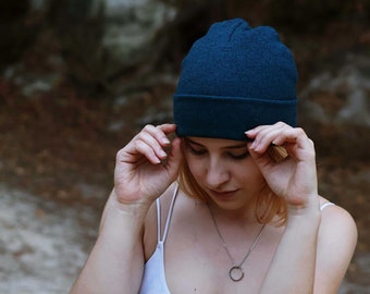 May include: A woman wearing a white tank top and a dark blue beanie. She is holding the beanie with both hands and looking down. She is wearing a silver necklace with a circular pendant.