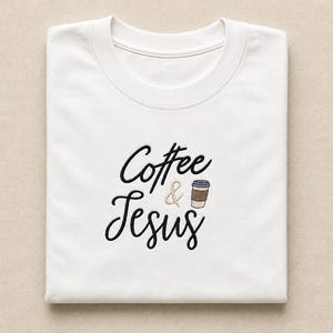 May include: A white crew neck t-shirt featuring black embroidered text that reads Coffee & Jesus. The ampersand is light brown, and a small embroidered graphic of a coffee cup with a brown sleeve and blue lid is positioned next to the text.