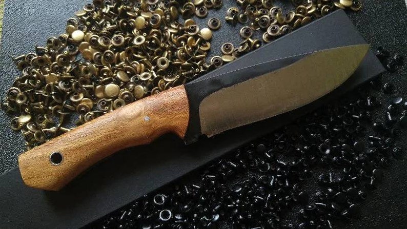 May include: A wooden handled knife with a black blade. The knife is lying on a black surface with a pile of gold and black metal fasteners.
