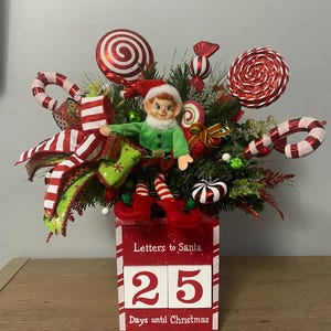 Puede incluir: Una decoración navideña festiva con una figura de elfo, bastones de caramelo y piruletas. La pieza central es una caja roja con el texto "Letters to Santa" y una pantalla que muestra "25 días para Navidad". El arreglo incluye follaje verde y cintas rojas y blancas.