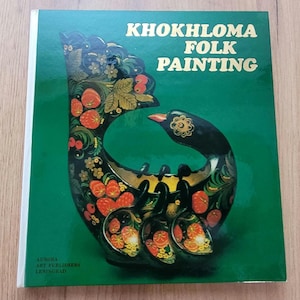 Könnte beinhalten: Ein Buch mit dem Titel "Khokhloma Folk Painting" mit grünem Einband. Das Cover zeigt ein schwarzes, verziertes, vogelähnliches Design mit roten Beeren und floralen Akzenten. Herausgegeben von Aurora Art Publishers, Leningrad.