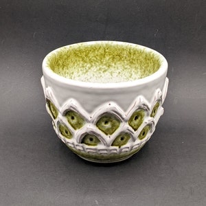 Puede incluir: Un tazón de cerámica blanco con un esmalte verde y un diseño texturizado. El tazón tiene un borde festoneado y un patrón de círculos superpuestos.