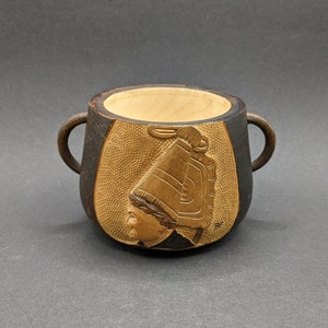 Può includere: Una tazza di legno con un design intagliato di una persona con un cappello. La tazza è marrone con un design intagliato marrone chiaro.
