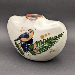 May include: Cream-colored ceramic vase with a bird and floral design. The bird has blue, orange, and black details, perched on a green leaf and blue flowers. The vase has a small opening at the top and is approximately 6 inches wide.
