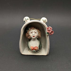 May include: A ceramic wall-hanging sculpture featuring a bust of a person with brown hair inside a white arched frame. The figure wears a green garment with two red circular accents. A pink rose is attached to the right side, and two small white spheres with green leaf patterns sit on top.