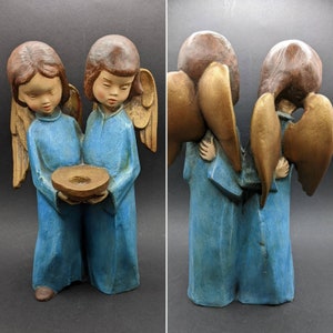 May include: Two blue angel figurines with wings and halos, standing side-by-side. The angel on the left is holding a small, round dish. Both angels have brown hair and painted facial features.