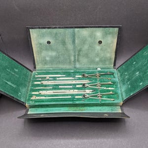 May include: A vintage drafting set in a black case with a green velvet interior. The set contains silver-coloured compasses, dividers, and other drawing tools, neatly arranged in fitted slots within the case.