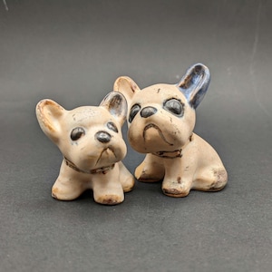 Può includere: Due piccole statuette di bulldog francese in ceramica con smalto beige chiaro e accenti marrone scuro su orecchie, occhi, naso e collari. I cani sono raffigurati in posizione seduta con orecchie dritte e tratti del viso ben definiti.