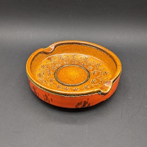 May include: A round orange and brown ceramic ashtray with a floral design. The ashtray has a wide rim and a small indentation in the centre.