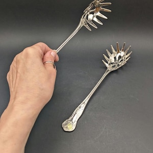 Op de afbeelding: Twee zilverkleurige spaghettilepels met een uniek ontwerp. De lepels hebben een lange steel en een kop met meerdere tanden. Eén lepel wordt door een hand vastgehouden. De lepels staan op een donkere achtergrond.
