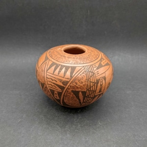 May include: A small, round, terracotta-colored clay pot with a wide, circular opening at the top. The exterior surface features a textured, mottled finish and is decorated with black geometric patterns, including triangles, lines, and shapes.