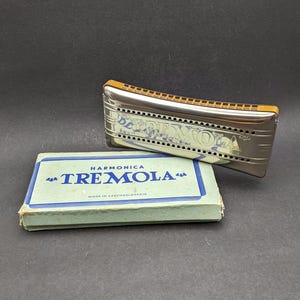 Pode incluir: Uma harmónica Tremola de metal prateado com pente castanho, apresentando o texto "TREMOLA" gravado na lateral. É acompanhada por uma caixa de cartão azul-claro com o texto "HARMONICA TREMOLA" e "MADE IN CZECHOSLOVAKIA" impresso.