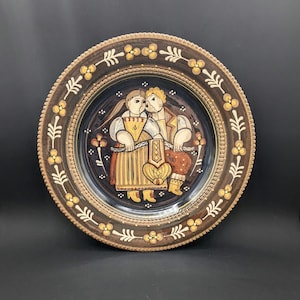 May include: A round ceramic plate with a dark blue background and a painted scene of two figures in traditional clothing. The figures are holding hands and looking at each other. The plate has a brown rim with a decorative border of white leaves and yellow flowers.