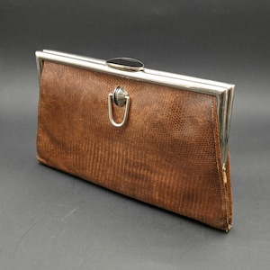 May include: A rectangular brown clutch bag with a textured lizard-skin pattern. The bag features a silver-toned metal frame, a top clasp closure with a black accent, and a decorative silver-toned U-shaped ring ornament centred on the front panel.