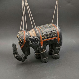 Könnte beinhalten: Eine schwarze Holz-Elefanten-Marionette mit aufwendigen Details. Der Elefant hat einen dekorativen Sattel mit roten und gelben Akzenten. Die Marionette ist an Schnüren aufgehängt, was darauf hindeutet, dass sie zum Spielen oder Ausstellen bestimmt ist. Der Rüssel des Elefanten ist beweglich.