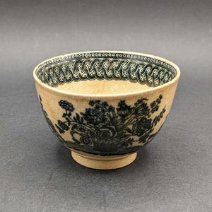May include: A cream-colored ceramic bowl with a dark teal floral design. The bowl features a decorative border and a pedestal base. The floral pattern includes roses and other flowers. The bowl is likely vintage or antique.